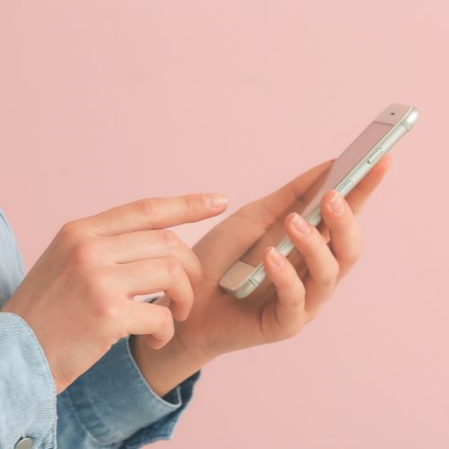 Pessoa segurando um celular com as duas mãos, digitando na tela sobre um fundo rosa claro.