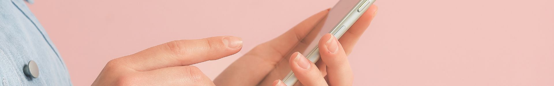 Pessoa segurando um celular com as duas mãos, digitando na tela sobre um fundo rosa claro.