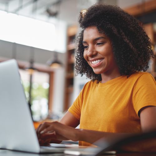 Mulher sorridente usa um notebook em ambiente moderno, vestindo camiseta amarela, representando trabalho ou estudo online.