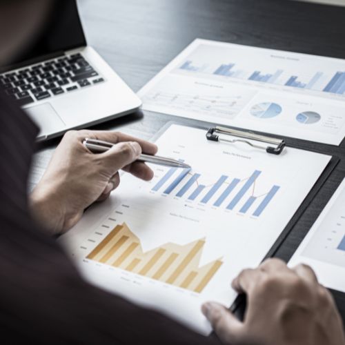 Mãos masculinas analisam relatórios impressos com gráficos e tabelas financeiras, ao lado de um notebook sobre a mesa de escritório.