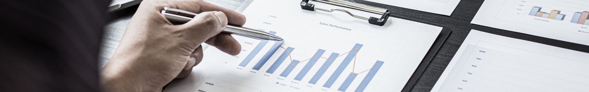 Mãos masculinas analisam relatórios impressos com gráficos e tabelas financeiras, ao lado de um notebook sobre a mesa de escritório.