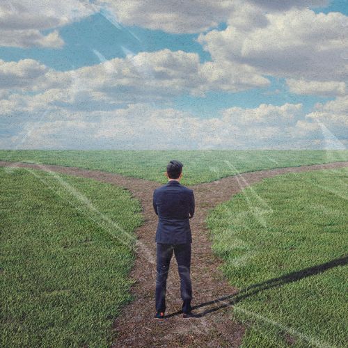 Homem de terno em encruzilhada em campo aberto, observando dois caminhos, representando decisão e escolha de direção.