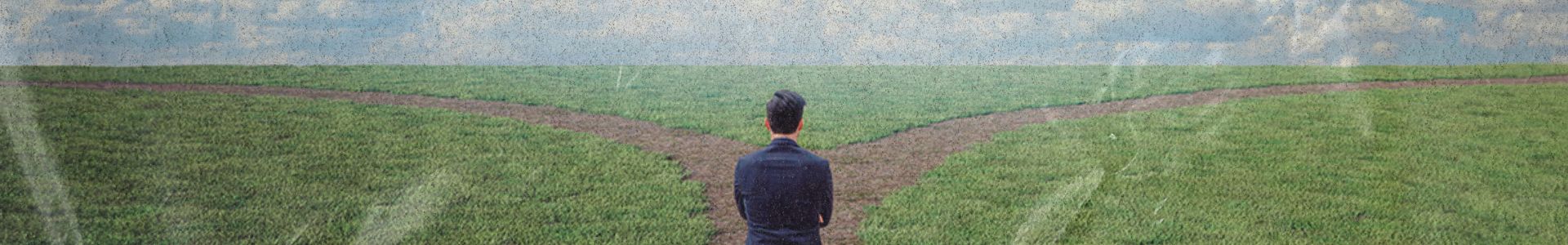 Homem de terno em encruzilhada em campo aberto, observando dois caminhos, representando decisão e escolha de direção.